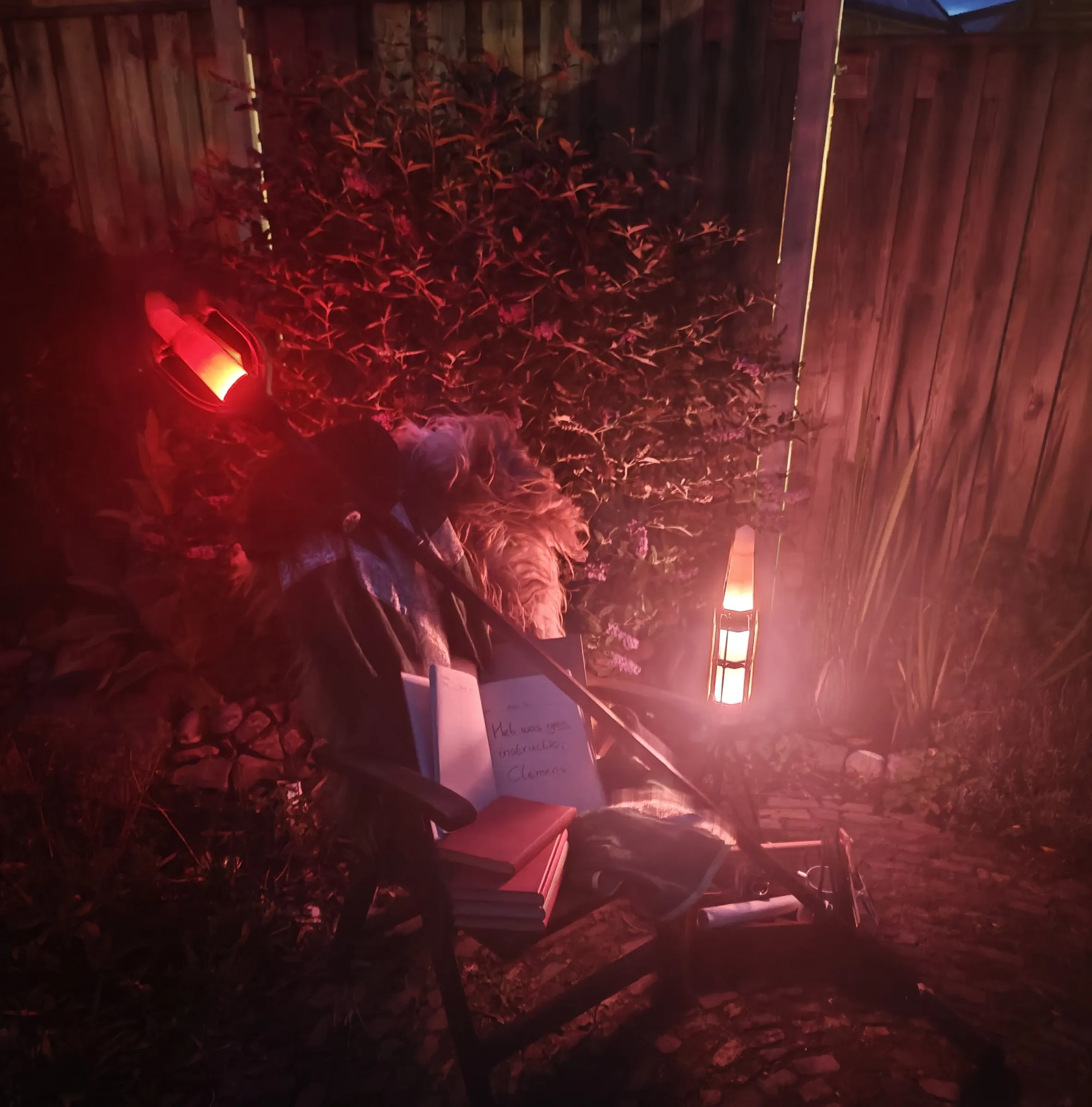 At night in the garden, a lantern glowing next to a chair with a fur, books and a lit staff